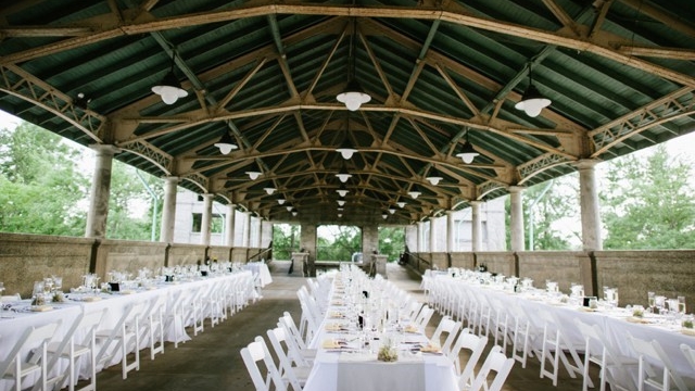 Outdoor wedding reception venue with long tables and white chairs under a green and wood pavilion.